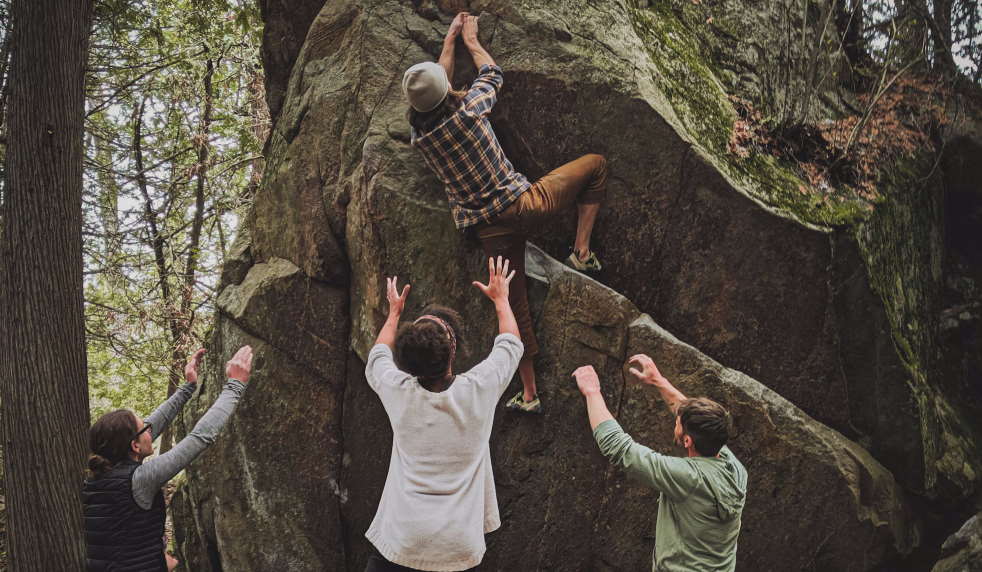 Person climbing large rock with three people standing below them with their arms outstretched.
