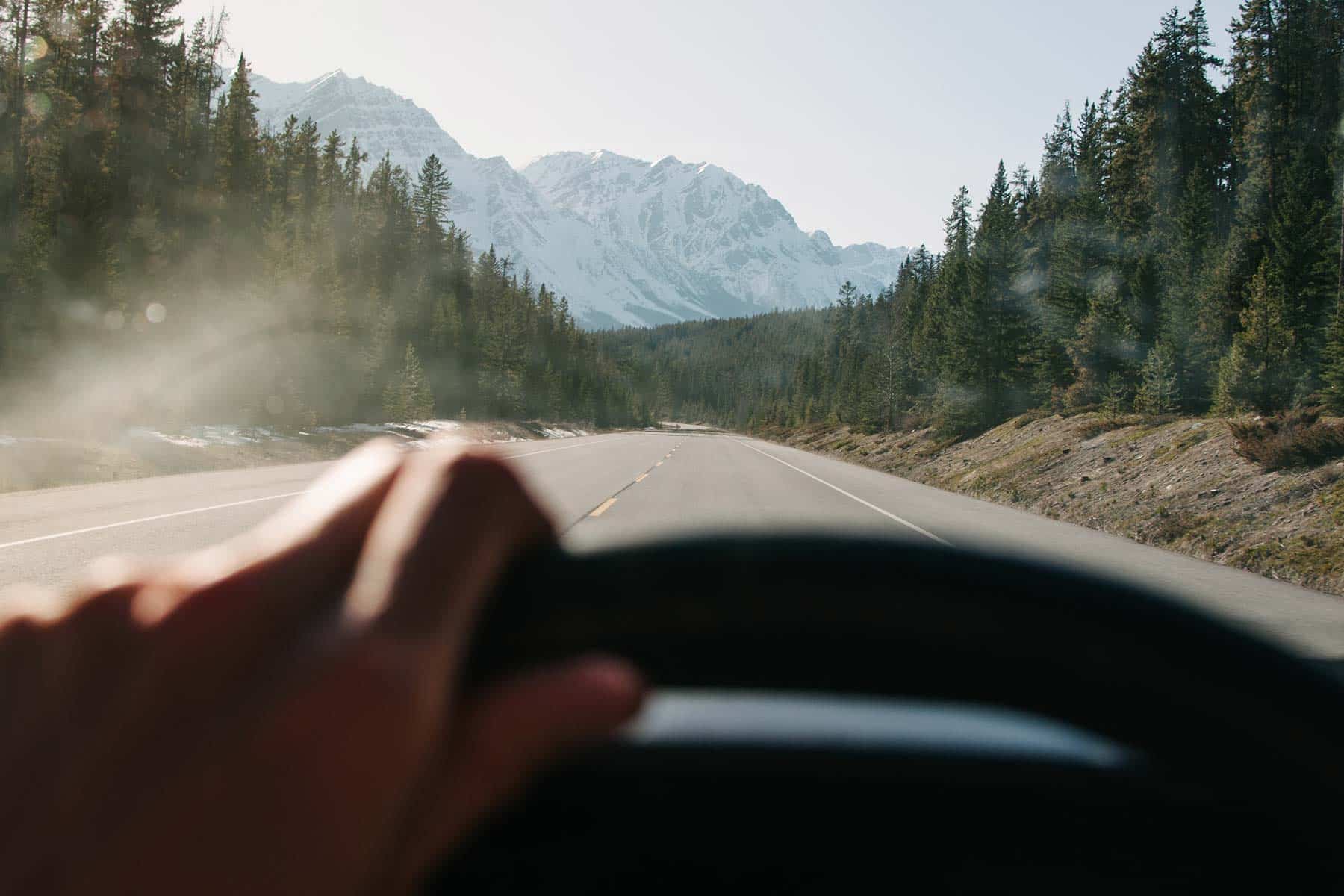 A person's hand on a steering wheel with a mountain road visible through the windshield